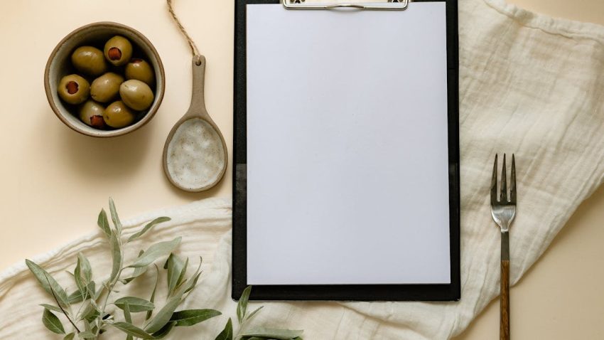 Flat lay of olives, a clipboard, and kitchen items creating a minimalist scene.